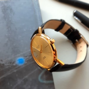 May include: A gold-tone wristwatch with a black leather strap. The watch face is round and has a gold-tone bezel. The watch face has a white background with black Roman numeral hour markers and black hands. The watch face also has the text "Poljot M1-CZK" and "23 Jewels Automatic" printed on it.