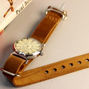 Puede incluir: Un reloj de pulsera de estilo vintage con una esfera blanca y n&uacute;meros negros. El reloj tiene una correa de cuero marr&oacute;n con costuras y un cierre de hebilla.