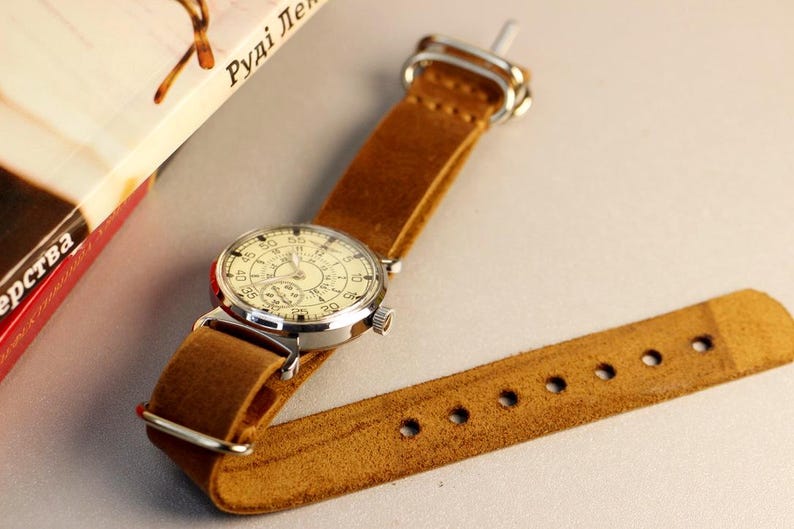 Puede incluir: Un reloj de pulsera de estilo vintage con una esfera blanca y n&uacute;meros negros. El reloj tiene una correa de cuero marr&oacute;n con costuras y un cierre de hebilla.