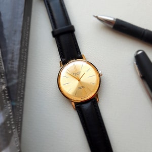 May include: A gold-tone wristwatch with a black leather strap. The watch face has a gold background with black markings and the words "Poljot De Luxe" and "23 Jewels" printed on it.