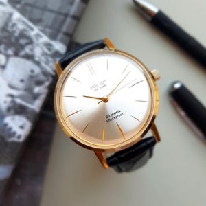 K&ouml;nnte beinhalten: Eine goldfarbene Armbanduhr mit wei&szlig;em Zifferblatt und schwarzem Lederarmband. Das Zifferblatt der Uhr hat schwarze r&ouml;mische Ziffern als Stundenmarkierungen und schwarze Zeiger. Das Zifferblatt der Uhr tr&auml;gt au&szlig;erdem den Text "Poljot de Luxe" und "23 jewels shockproof".