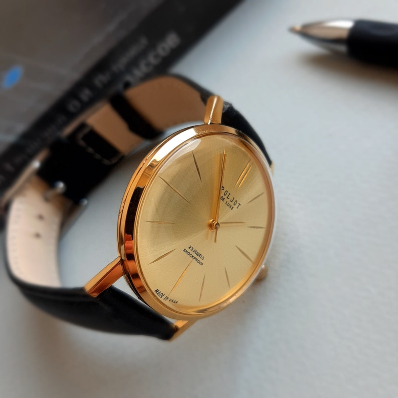 May include: A gold-tone wristwatch with a black leather strap. The watch face is gold with black markings and the text "Poljot De Luxe" and "23 Jewels Shockproof" printed on it. The watch is made in the USSR.
