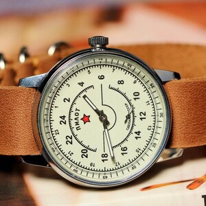 Puede incluir: Un reloj de pulsera plateado con una esfera blanca y agujas negras. El reloj tiene una correa de cuero marr&oacute;n y una estrella roja en la esfera. La esfera del reloj tiene la palabra "ПИЛОТ" impresa en ella.