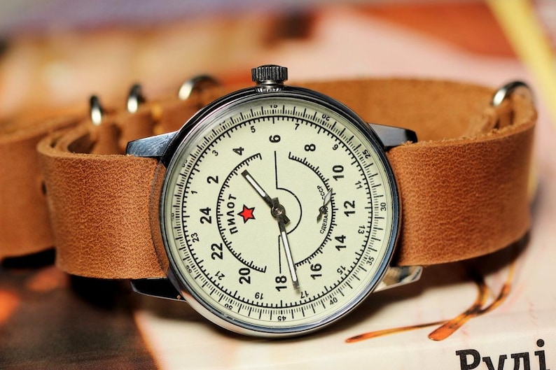 Puede incluir: Un reloj de pulsera plateado con una esfera blanca y agujas negras. El reloj tiene una correa de cuero marr&oacute;n y una estrella roja en la esfera. La esfera del reloj tiene la palabra "ПИЛОТ" impresa en ella.