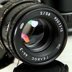 Puede incluir: Lente de c&aacute;mara negra con el texto "2/58" y "9003315" en blanco. La lente tiene un anillo de enfoque estriado y un elemento de vidrio reflectante. Otras partes de la c&aacute;mara son visibles en el fondo.