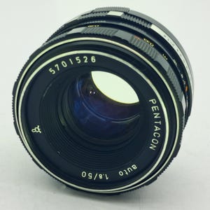 May include: A black Pentacon auto 1.8/50 camera lens with silver accents. The lens has a series of rings for focus and aperture control. The lens has the number 5701526 printed on it.