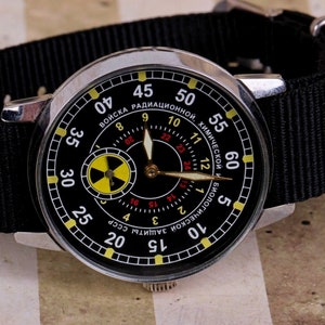 May include: A black wristwatch with a silver case and a black nylon strap. The watch face has a black background with white and yellow markings. The watch face also has a small yellow and black radiation symbol. The watch face has the text "ВОЙСКА РАДИАЦИОННОЙ, ХИМИЧЕСКОЙ И БИОЛОГИЧЕСКОЙ ЗАЩИТЫ СССР" in Cyrillic.