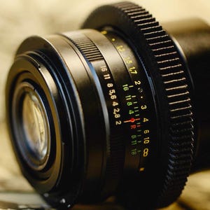 May include: A black camera lens with a silver aperture ring and a depth of field scale marked in f-stops from 2.8 to 16. The lens has a gear ring for attaching to a camera.