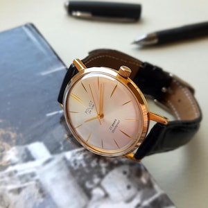 K&ouml;nnte beinhalten: Eine goldfarbene Armbanduhr mit wei&szlig;em Zifferblatt und schwarzem Lederarmband. Das Zifferblatt der Uhr hat goldfarbene Zeiger und Markierungen, und der Text "POLJOT de Luxe" und "23 jewels shockproof" ist auf dem Zifferblatt aufgedruckt.