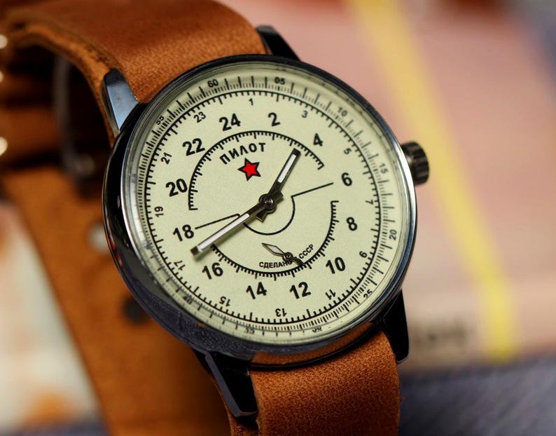 Puede incluir: Un reloj de pulsera vintage con una correa de cuero marr&oacute;n y una esfera blanca. La esfera tiene marcas negras y una estrella roja con la palabra "ПИЛОТ" impresa en ella. El reloj tambi&eacute;n tiene el texto "СДЕЛАНО В СССР" impreso en la esfera.