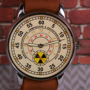 May include: A silver wristwatch with a white face and black hands. The face has a yellow and black radiation symbol in the center and the text "ВОЙСКА РАДИАЦИОННОЙ, ХИМИЧЕСКОЙ И БИОЛОГИЧЕСКОЙ ЗАЩИТЫ СССР" around the outside.