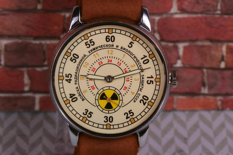 May include: A silver wristwatch with a white face and black hands. The face has a yellow and black radiation symbol in the center and the text "ВОЙСКА РАДИАЦИОННОЙ, ХИМИЧЕСКОЙ И БИОЛОГИЧЕСКОЙ ЗАЩИТЫ СССР" around the outside.