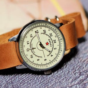 Puede incluir: Un reloj de pulsera vintage con una esfera blanca y agujas negras. El reloj tiene una correa de cuero marr&oacute;n y una estrella roja en la esfera con la palabra "Пилот" impresa debajo.