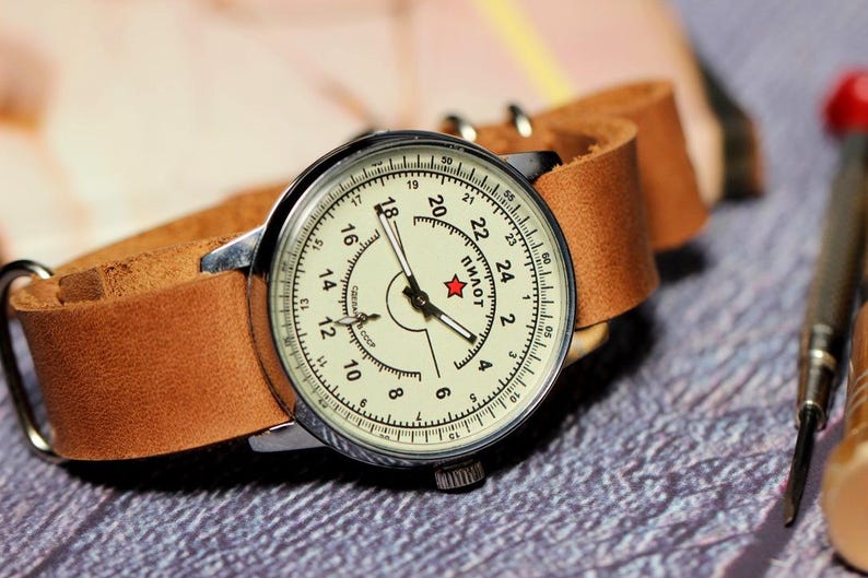 Puede incluir: Un reloj de pulsera vintage con una esfera blanca y agujas negras. El reloj tiene una correa de cuero marr&oacute;n y una estrella roja en la esfera con la palabra "Пилот" impresa debajo.