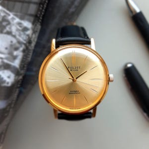 May include: A gold-tone wristwatch with a black leather strap. The watch face is gold with black markings and the text "Poljot De Luxe 23 Jewels Shockproof Made in USSR".