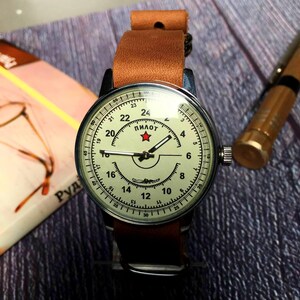 Puede incluir: Un reloj de pulsera vintage con una correa de cuero marr&oacute;n. La esfera del reloj es blanca con marcas negras y una estrella roja. El texto "ПИЛОТ" est&aacute; impreso en la esfera del reloj.