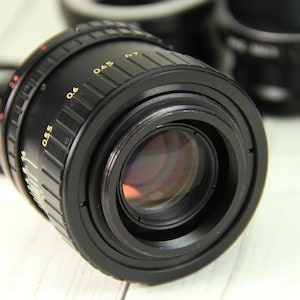 Puede incluir: Un objetivo de c&aacute;mara negro con marcas doradas, incluyendo n&uacute;meros y medidas. El objetivo tiene un elemento de vidrio reflectante y se encuentra sobre un fondo borroso de otros accesorios de c&aacute;mara. El objetivo est&aacute; dise&ntilde;ado para fotograf&iacute;a.