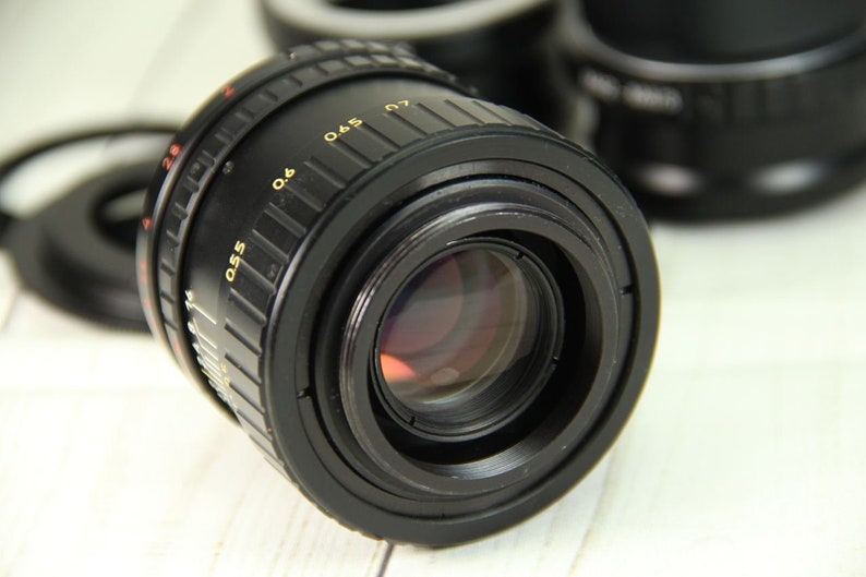 Puede incluir: Un objetivo de c&aacute;mara negro con marcas doradas, incluyendo n&uacute;meros y medidas. El objetivo tiene un elemento de vidrio reflectante y se encuentra sobre un fondo borroso de otros accesorios de c&aacute;mara. El objetivo est&aacute; dise&ntilde;ado para fotograf&iacute;a.