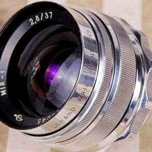 May include: A vintage MIR-1 2.8/37 camera lens with a silver and black finish. The lens has the text "GRAND PRIX BRUSSELS" and other markings. The lens is designed for photography and has a reflective purple interior.