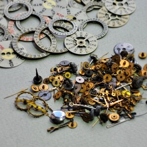 May include: A collection of watch parts including numerous circular date wheels with printed numbers and Cyrillic text, alongside a dense pile of small brass and steel mechanical gears, pinions, and watch hands scattered across a flat surface.
