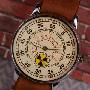 May include: A silver wristwatch with a white face and black hands. The watch face has a yellow and black radiation symbol in the center and the text "ВОЙСКА РАДИАЦИОННОЙ, ХИМИЧЕСКОЙ И БИОЛОГИЧЕСКОЙ ЗАЩИТЫ СССР" around the outside.
