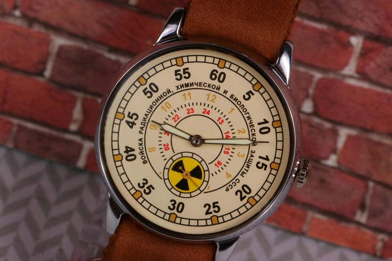May include: A silver wristwatch with a white face and black hands. The watch face has a yellow and black radiation symbol in the center and the text "ВОЙСКА РАДИАЦИОННОЙ, ХИМИЧЕСКОЙ И БИОЛОГИЧЕСКОЙ ЗАЩИТЫ СССР" around the outside.