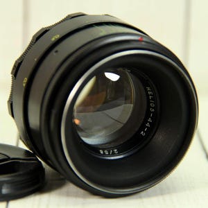 May include: A black camera lens with a silver front ring and a detached black lens cap. The lens barrel features the text HELIOS-44-2 and 2/58 printed in white. The design includes a textured focus ring and a red alignment dot on the top edge.