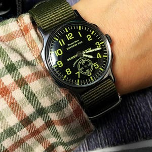 Puede incluir: Un reloj de pulsera negro y verde con una esfera redonda y una correa de nailon verde. La esfera del reloj tiene marcas amarillas y el texto "Тадж-Бек ШТОРМ 333 ЗЕНИТ СПЕЦНАЗ 37 DOM" en cirílico.
