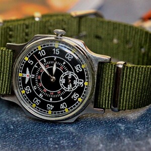 May include: A black and silver wristwatch with a green nylon strap. The watch face has a black background with white numbers and a small second hand dial. The watch has a red airplane symbol on the face.