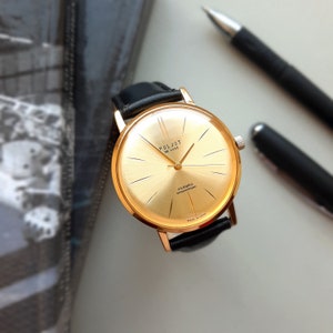 May include: A gold-tone wristwatch with a black leather strap. The watch face is gold with black markings and the text "Poljot De Luxe 23 Jewels Shockproof Made in USSR".