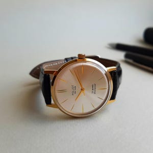 K&ouml;nnte beinhalten: Eine goldfarbene Armbanduhr mit einem schwarzen Lederarmband. Das Zifferblatt der Uhr ist wei&szlig; mit goldfarbenen Zeigern und Markierungen. Das Zifferblatt der Uhr tr&auml;gt au&szlig;erdem den Text "Poljot de Luxe" und "23 jewels shockproof".