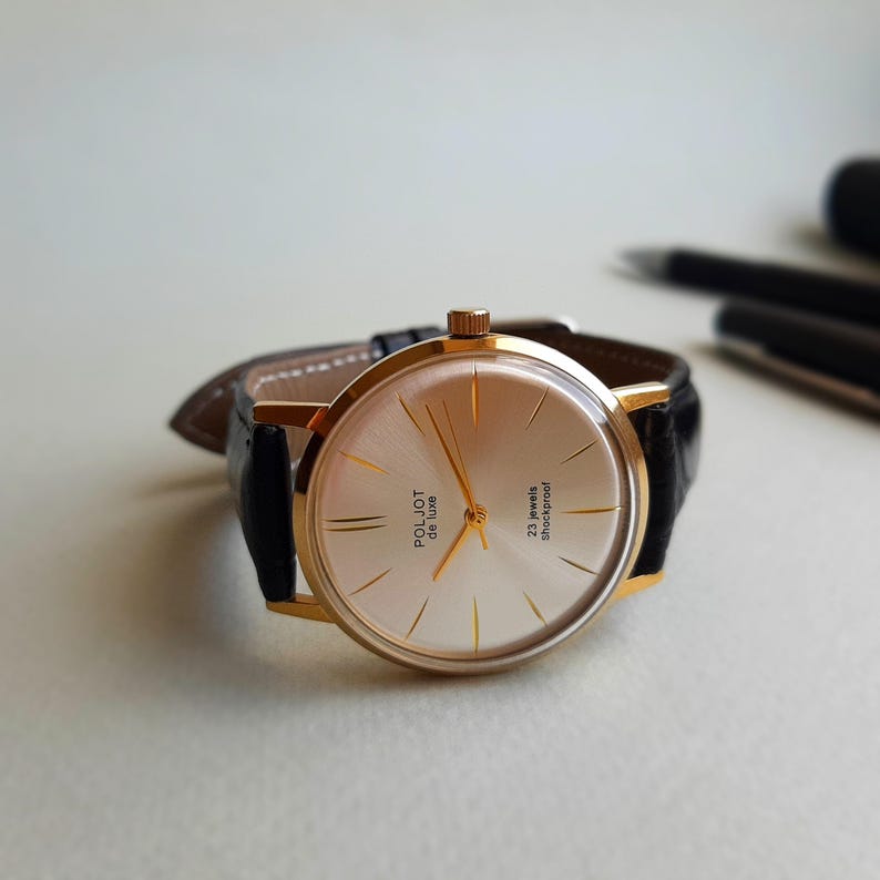 K&ouml;nnte beinhalten: Eine goldfarbene Armbanduhr mit einem schwarzen Lederarmband. Das Zifferblatt der Uhr ist wei&szlig; mit goldfarbenen Zeigern und Markierungen. Das Zifferblatt der Uhr tr&auml;gt au&szlig;erdem den Text "Poljot de Luxe" und "23 jewels shockproof".