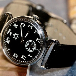 Puede incluir: Un reloj de pulsera negro con una caja plateada y una correa de cuero negro. La esfera del reloj tiene números hebreos y una estrella de David.