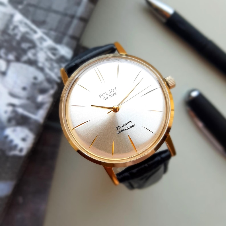 K&ouml;nnte beinhalten: Eine goldfarbene Armbanduhr mit wei&szlig;em Zifferblatt und schwarzem Lederarmband. Das Zifferblatt der Uhr hat schwarze r&ouml;mische Ziffern als Stundenmarkierungen und schwarze Zeiger. Das Zifferblatt der Uhr tr&auml;gt au&szlig;erdem den Text "Poljot de Luxe" und "23 jewels shockproof".