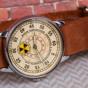May include: A vintage wristwatch with a white face and black hands. The watch face has a yellow and black radiation symbol in the center and the text "ВОЙСКА РАДИАЦИОННОЙ, ХИМИЧЕСКОЙ И БИОЛОГИЧЕСКОЙ ЗАЩИТЫ СССР" around the outside.