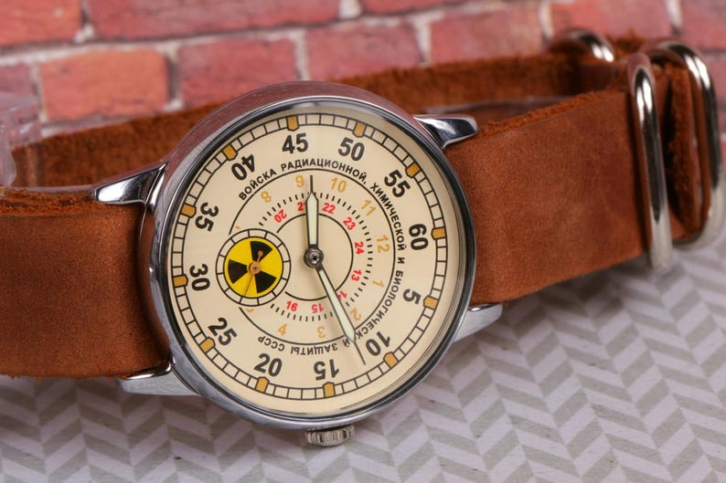 May include: A vintage wristwatch with a white face and black hands. The watch face has a yellow and black radiation symbol in the center and the text "ВОЙСКА РАДИАЦИОННОЙ, ХИМИЧЕСКОЙ И БИОЛОГИЧЕСКОЙ ЗАЩИТЫ СССР" around the outside.