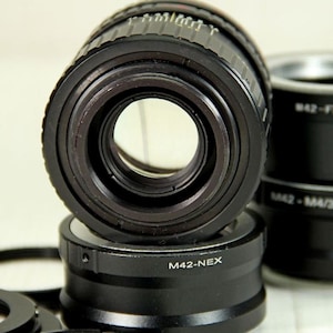 Puede incluir: Primer plano de varios adaptadores de lentes de c&aacute;mara negros. El adaptador de lente principal est&aacute; enfocado, con una gran apertura central. Los adaptadores est&aacute;n etiquetados con texto que incluye "M42-NEX" y "M42-FX".