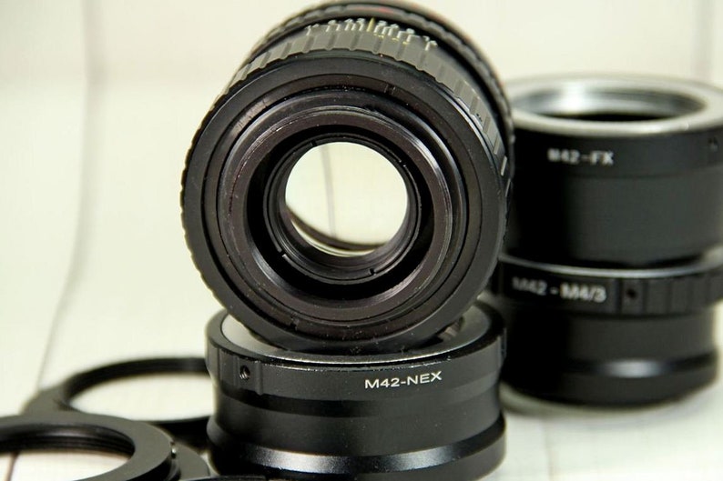 Puede incluir: Primer plano de varios adaptadores de lentes de c&aacute;mara negros. El adaptador de lente principal est&aacute; enfocado, con una gran apertura central. Los adaptadores est&aacute;n etiquetados con texto que incluye "M42-NEX" y "M42-FX".