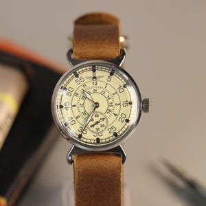Puede incluir: Un reloj de pulsera vintage con una esfera blanca y marcas negras. El reloj tiene una correa de cuero marr&oacute;n y una caja plateada. La esfera del reloj tiene un peque&ntilde;o dial de segundero y los n&uacute;meros 5, 10, 15, 20, 25, 30, 35, 40, 45, 50, 55 y 60 alrededor del borde exterior.