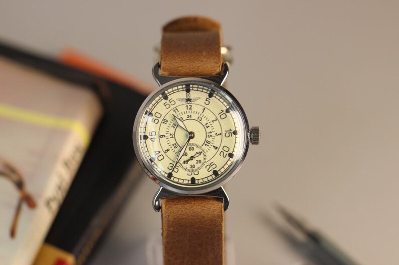 Puede incluir: Un reloj de pulsera vintage con una esfera blanca y marcas negras. El reloj tiene una correa de cuero marr&oacute;n y una caja plateada. La esfera del reloj tiene un peque&ntilde;o dial de segundero y los n&uacute;meros 5, 10, 15, 20, 25, 30, 35, 40, 45, 50, 55 y 60 alrededor del borde exterior.