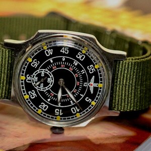 May include: A silver wristwatch with a black face and yellow markings. The watch has a green nylon strap and a small subdial at the 12 o'clock position. The watch face has the numbers 12, 1, 2, 3, 4, 5, 10, 15, 20, 25, 30, 35, 40, 45, 50, 55, and a small airplane symbol.