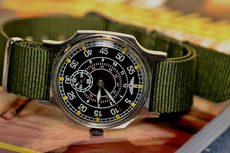 May include: A silver wristwatch with a black face and yellow markings. The watch has a green nylon strap and a small subdial at the 12 o'clock position. The watch face has the numbers 12, 1, 2, 3, 4, 5, 10, 15, 20, 25, 30, 35, 40, 45, 50, 55, and a small airplane symbol.
