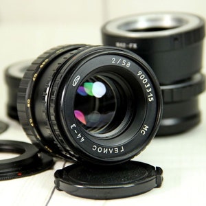 Puede incluir: Un objetivo de c&aacute;mara negro con el texto "2/58" y "9003315" es el punto focal. El objetivo est&aacute; rodeado de otros accesorios de c&aacute;mara negros, incluyendo una tapa de objetivo y anillos adaptadores. El objetivo tiene una superficie reflectante.