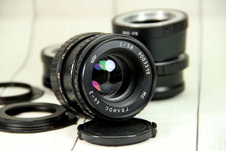 Puede incluir: Un objetivo de c&aacute;mara negro con el texto "2/58" y "9003315" es el punto focal. El objetivo est&aacute; rodeado de otros accesorios de c&aacute;mara negros, incluyendo una tapa de objetivo y anillos adaptadores. El objetivo tiene una superficie reflectante.