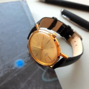 May include: A gold-tone wristwatch with a black leather strap. The watch face is round and has a gold-tone bezel. The watch face has black hands and markers. The watch face also has the text "Poljot de Luxe" and "23 Jewels shockproof" printed on it.