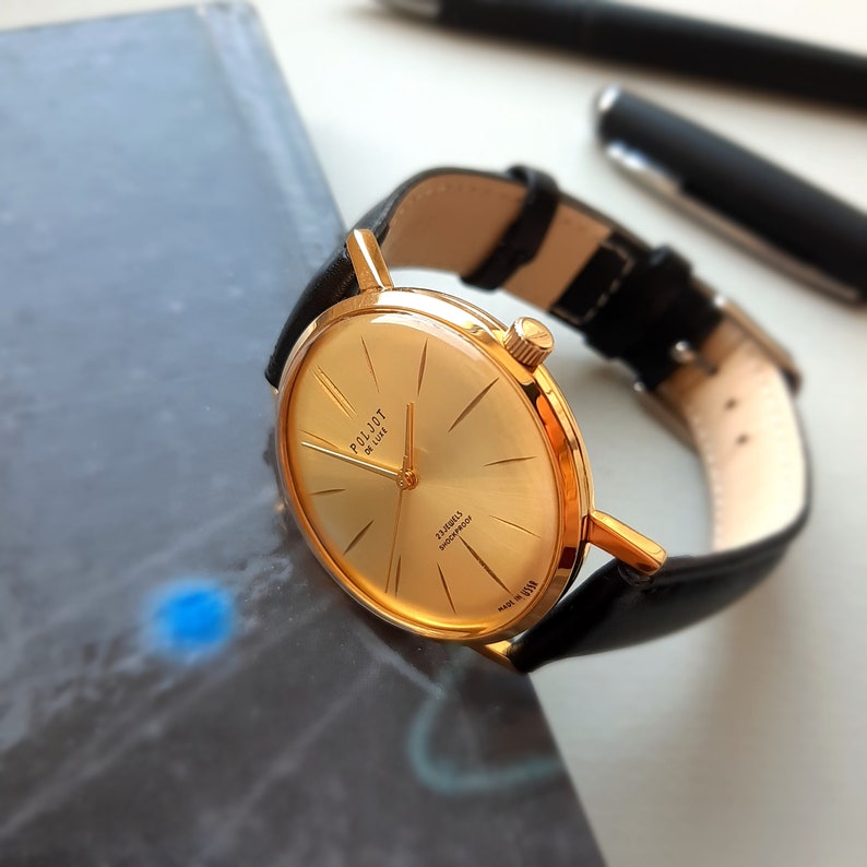 May include: A gold-tone wristwatch with a black leather strap. The watch face is round and has a gold-tone bezel. The watch face has black hands and markers. The watch face also has the text "Poljot de Luxe" and "23 Jewels shockproof" printed on it.