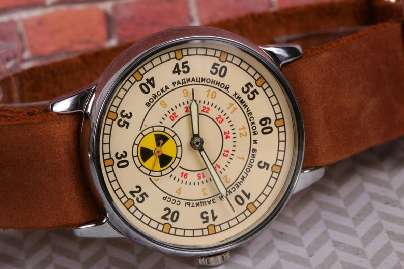 May include: A white wristwatch with a brown leather band. The watch face has a yellow and black radiation symbol and the text "ВОЙСКА РАДИАЦИОННОЙ. ХИМИЧЕСКОЙ И БИОЛОГИЧЕСКОЙ ЗАЩИТЫ СССР" in Russian.