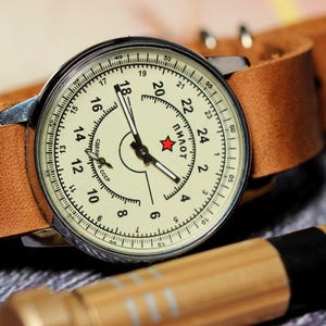 Puede incluir: Un reloj de pulsera plateado con una esfera blanca y agujas negras. El reloj tiene una correa de cuero marr&oacute;n y la esfera dice "ПИЛОТ" en letras cir&iacute;licas.
