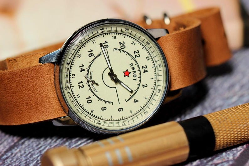 Puede incluir: Un reloj de pulsera plateado con una esfera blanca y agujas negras. El reloj tiene una correa de cuero marr&oacute;n y la esfera dice "ПИЛОТ" en letras cir&iacute;licas.