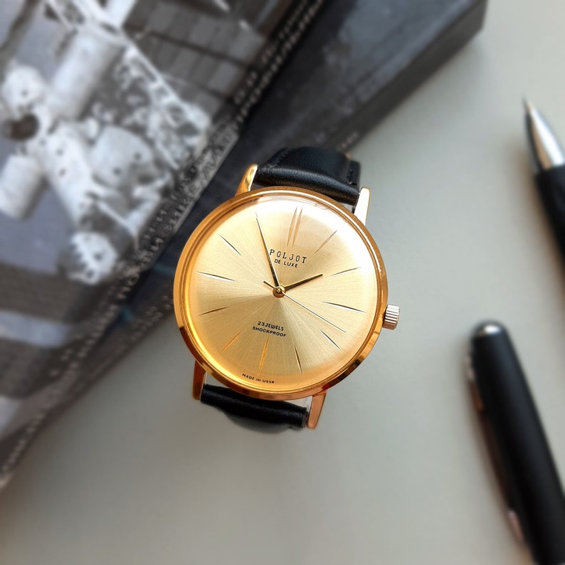 May include: A gold-tone wristwatch with a black leather strap. The watch face is gold with black markings and the words "Poljot De Luxe" and "23 Jewels Shockproof" printed on it.